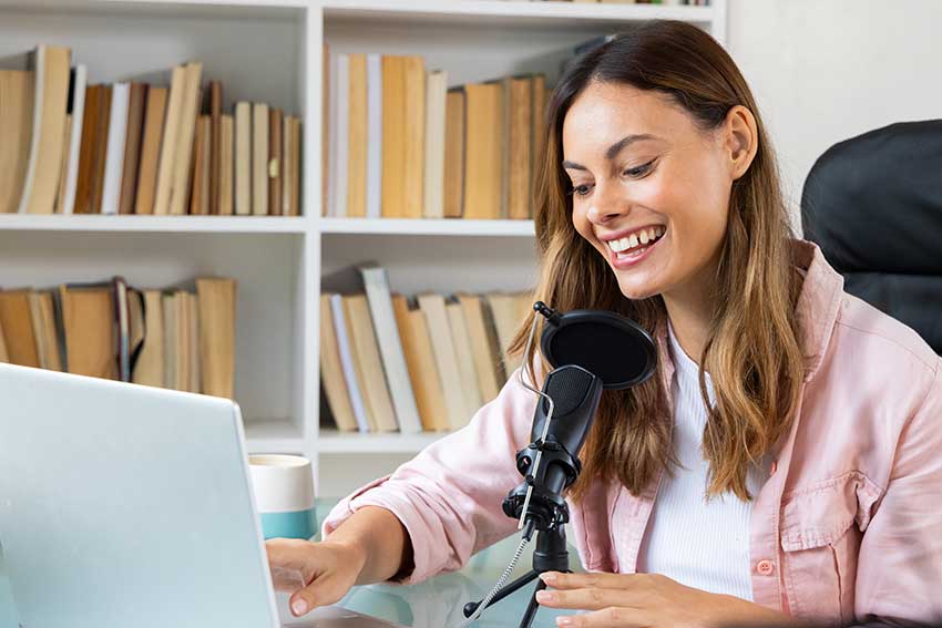 Woman recording podcast at home, smiling and using laptop with microphone.