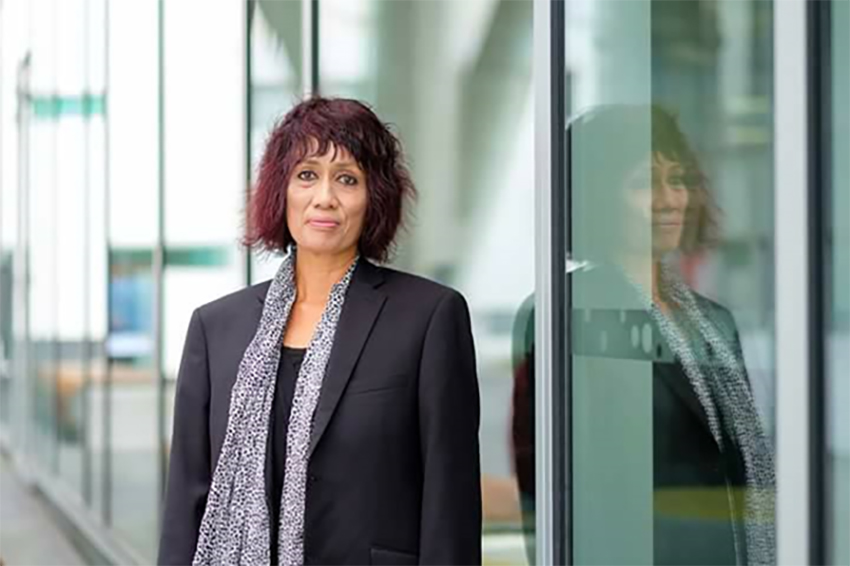 Associate Professor Teena Brown Pulu, slightly smiling and wearing a suit jacket and scarf, with her image reflected next to her in glass.